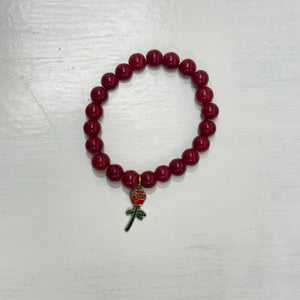Glossy deep-red beaded bracelet with a small red rose charm, photographed on a light wooden surface.