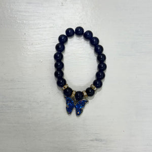 Dark beaded bracelet with gold-tone spacer beads and a sparkling blue butterfly charm, photographed on a light surface.